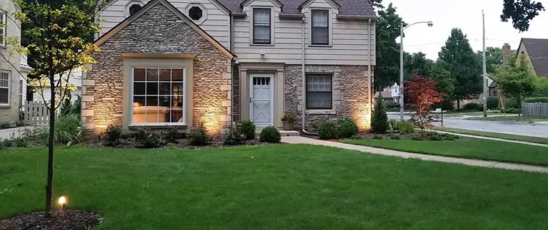 This image shows a two-story house with stone and siding exterior during dusk, featuring exterior lighting, well-maintained lawn, and a landscaped garden.