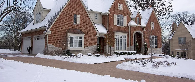 A large brick house with a snowy landscape. The driveway is cleared, and leafless trees suggest a cold winter setting. Snow blankets the ground and rooftops.