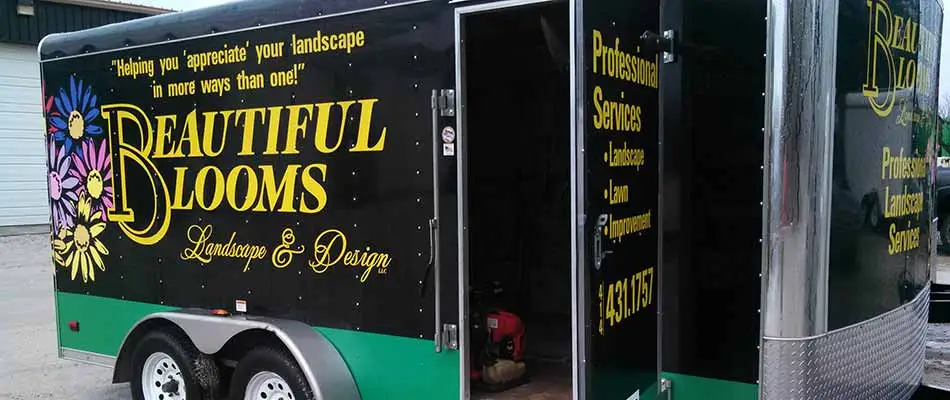 A black trailer with "Beautiful Blooms Landscape & Design" branding, colorful flower graphics, lists of services offered, and a contact number on its side.