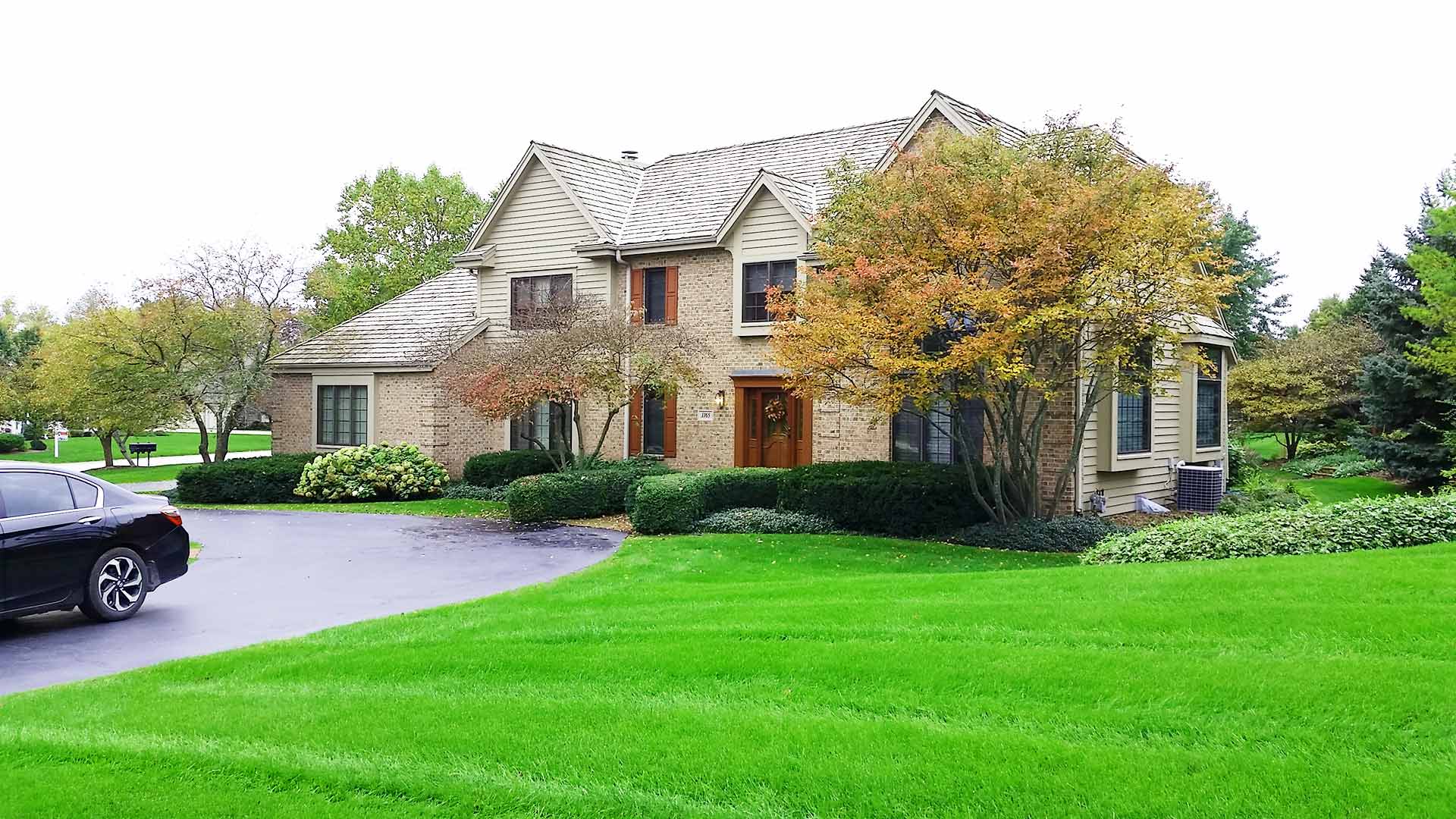 This is a suburban two-story house with beige siding, white trim, a well-kept lawn, a driveway with a black car, and autumn trees.