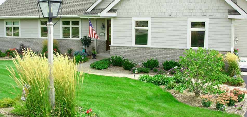 A single-story suburban home with a well-manicured lawn, landscaping, an American flag, and a street lamp in the foreground on a sunny day.