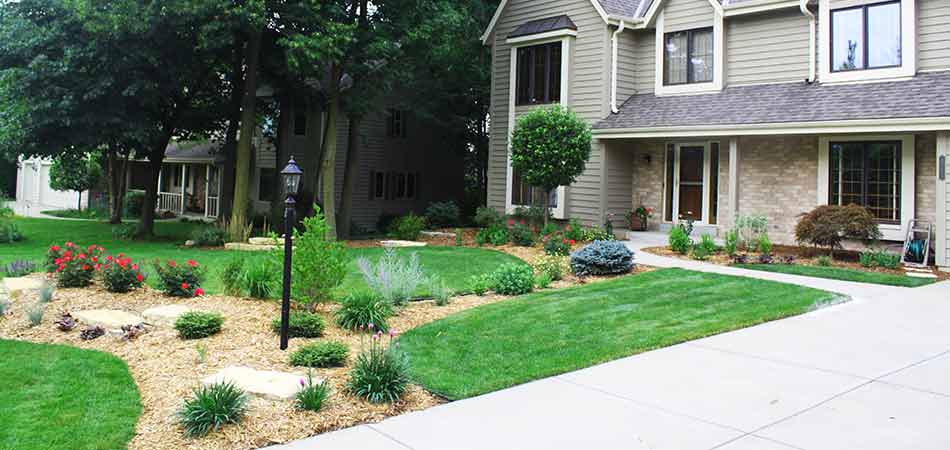 This is a suburban landscape with a two-story house, neatly trimmed lawn, landscaped garden, pathway, a street lamp, and tall trees.