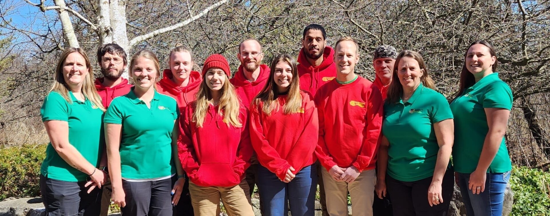 This image shows eleven people posing outdoors in matching red and green tops, likely colleagues, with a natural backdrop of trees and shrubbery.