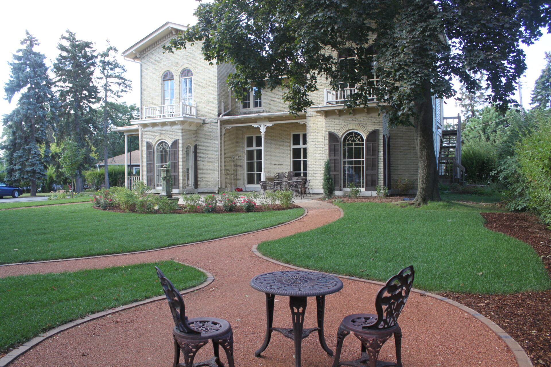 A two-story brick house with white trim, ornate balconies, and arched windows surrounded by a manicured garden, brick pathways, and a wrought-iron table set.