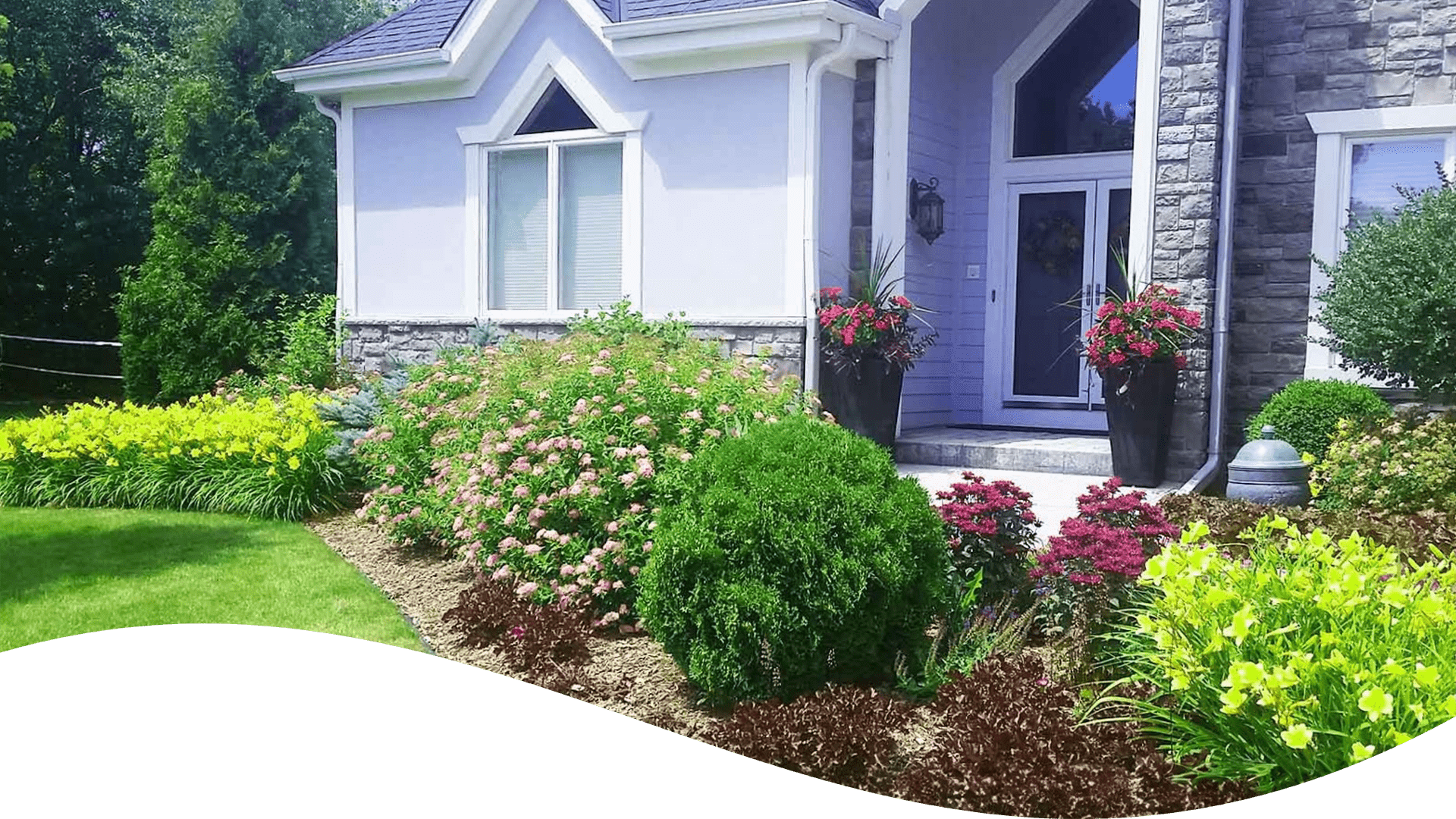 The image shows a well-maintained garden in front of a house with stone accents, colorful flowers, shrubs, and a neatly trimmed lawn.