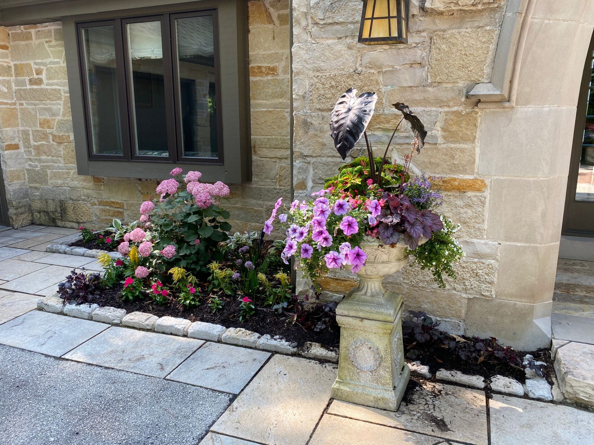A stone house with a flower bed containing hydrangeas and assorted plants, next to a window and an outdoor lamp, on a paved surface.
