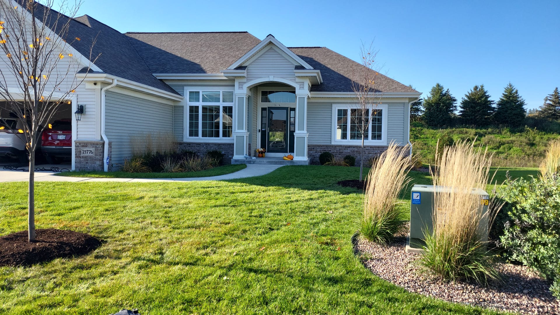 This image shows a single-story suburban home with beige siding, a garage, green lawn, and ornamental grasses, under a clear blue sky.