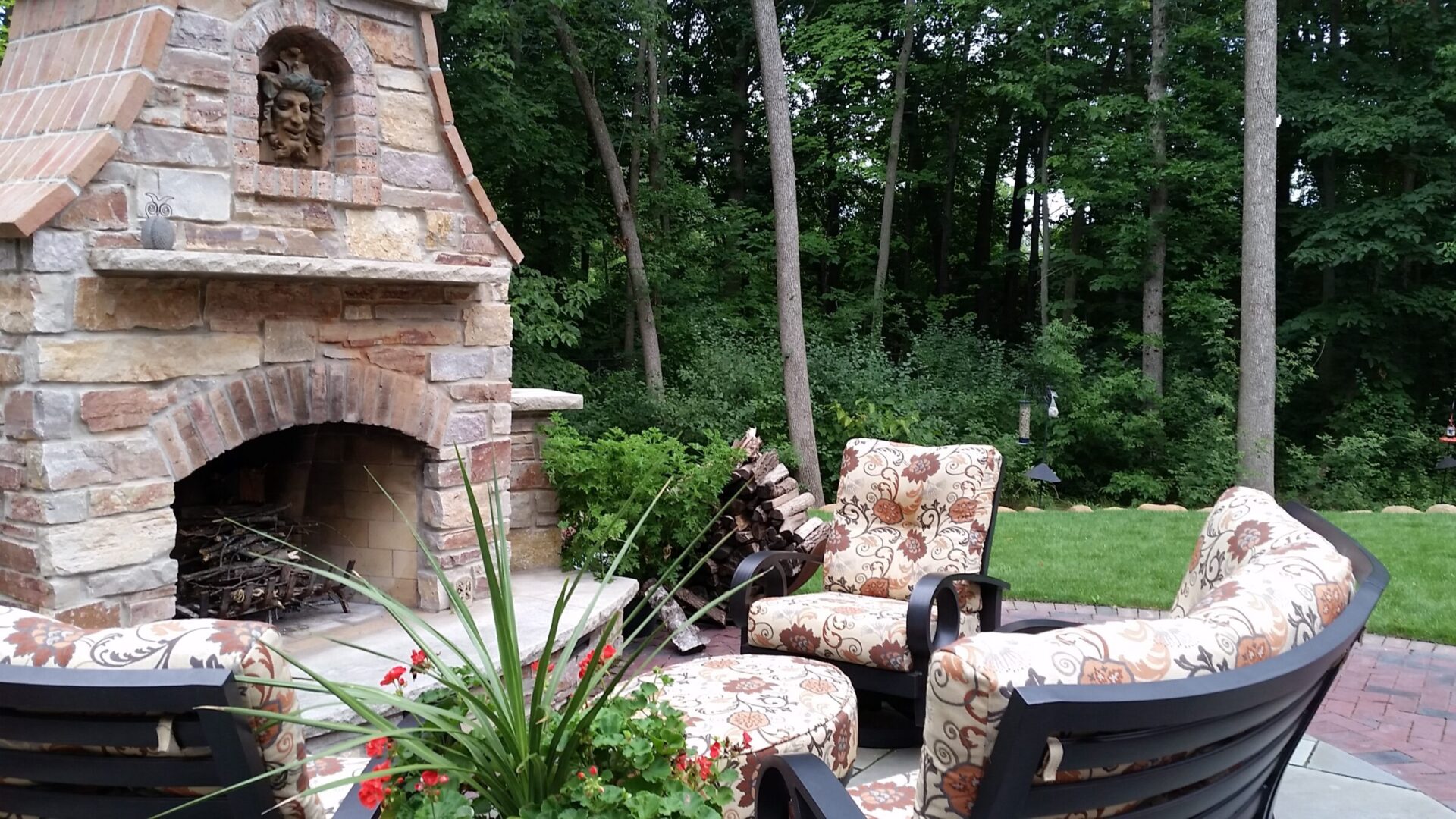 An outdoor stone fireplace with a wood pile is surrounded by patterned upholstered chairs. Lush greenery and trees create a tranquil backyard setting.