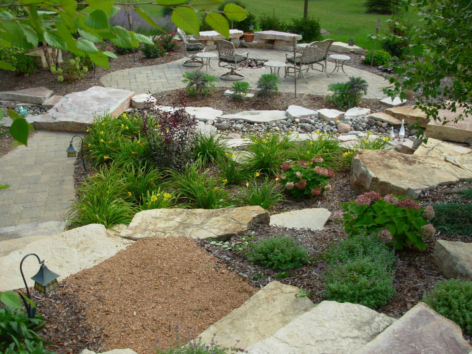 An inviting garden with paved paths, lush plants, decorative stones, outdoor seating, and lanterns, surrounded by natural landscaping elements.