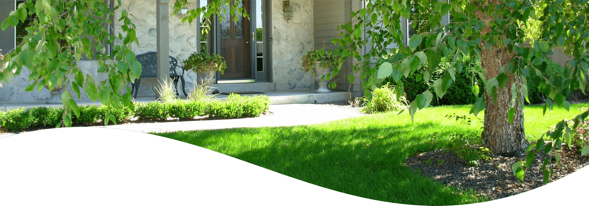 This image shows a well-manicured lawn with trees in front of a stone facade house featuring a dark wood door and wall-mounted lanterns.
