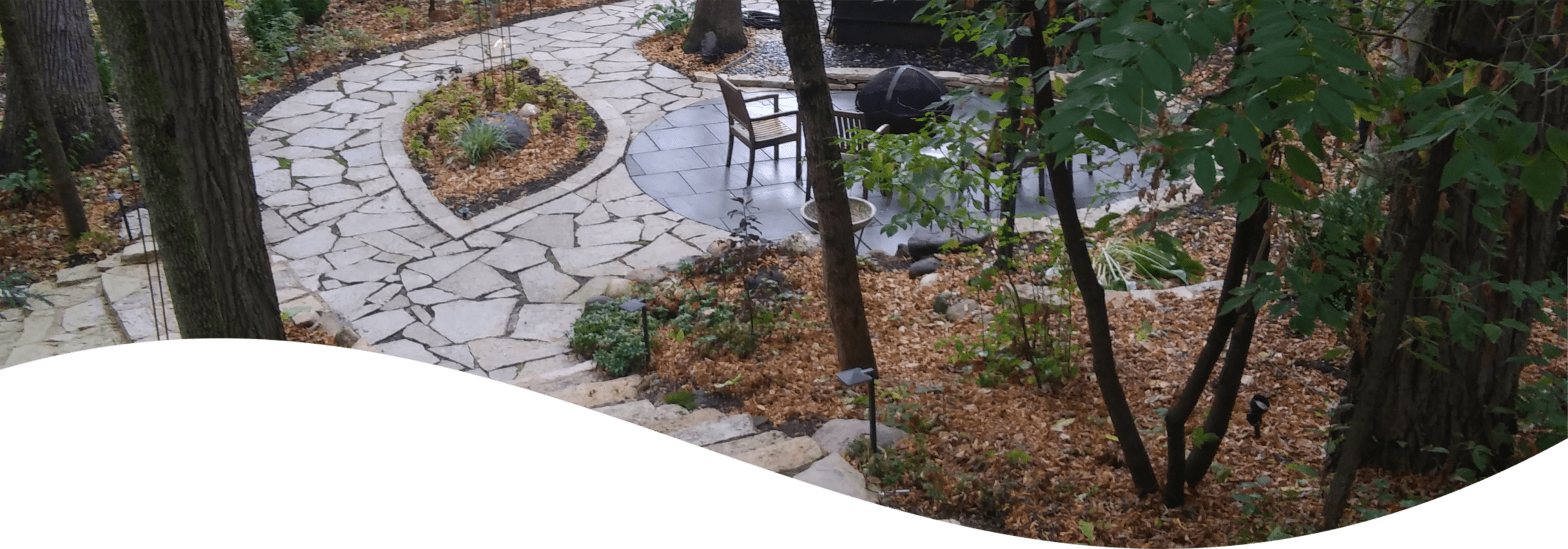A serene garden pathway meanders through trees and foliage, leading to a small patio area with a table and chairs, suggesting a tranquil outdoor space.