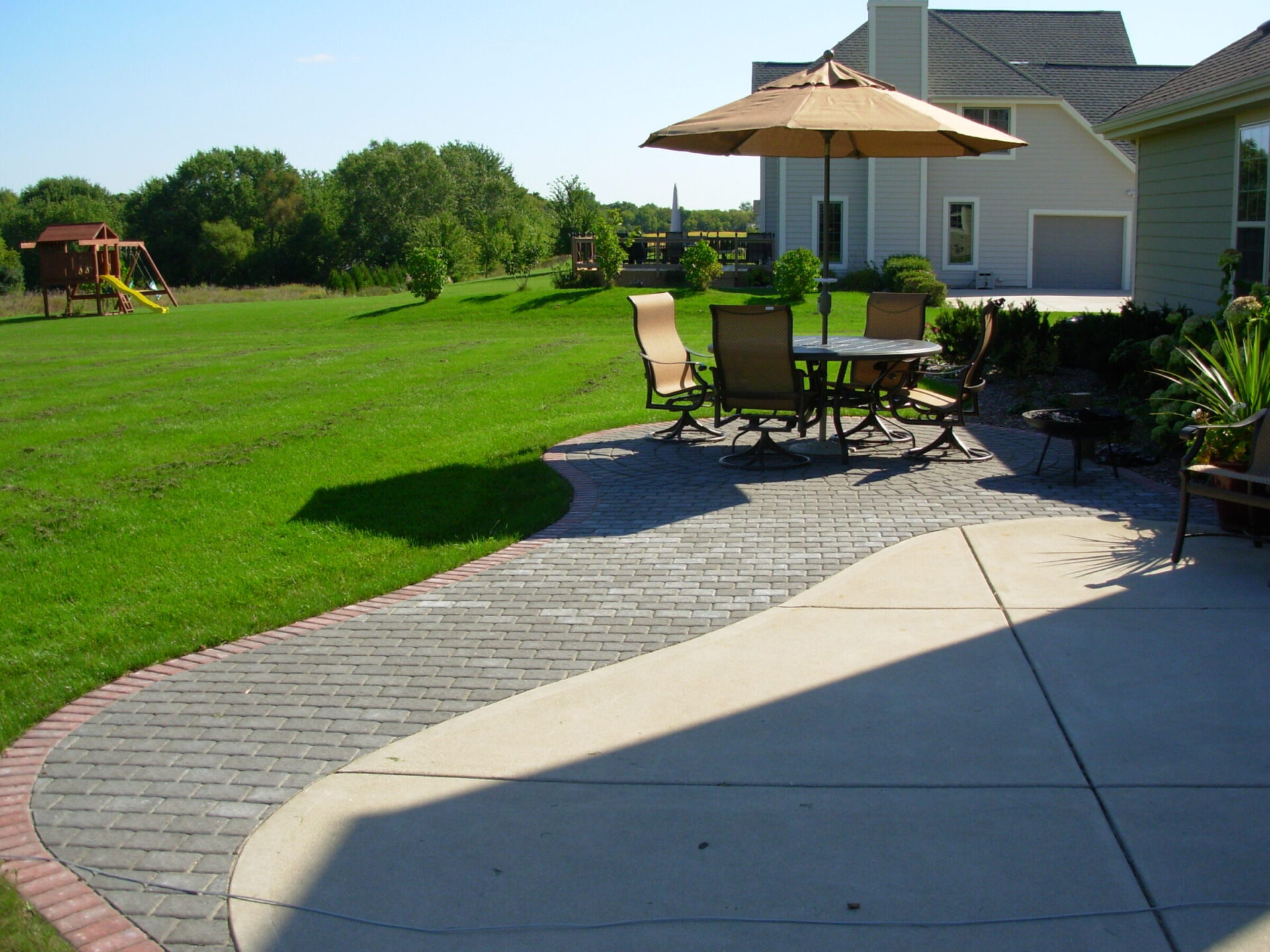 This image shows a backyard with a paved patio, outdoor dining set under an umbrella, a play structure, well-kept lawn, and a house.