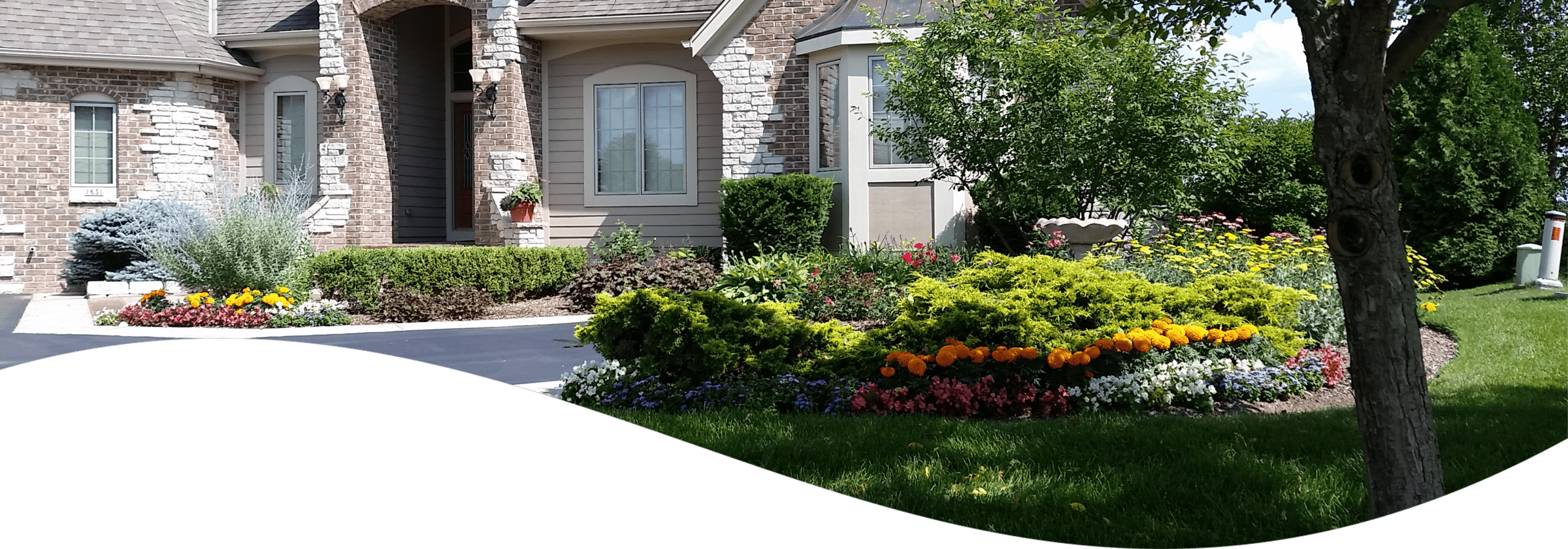 A suburban house with a stone and siding facade has a well-manicured lawn and colorful, blooming flower beds under a clear blue sky.