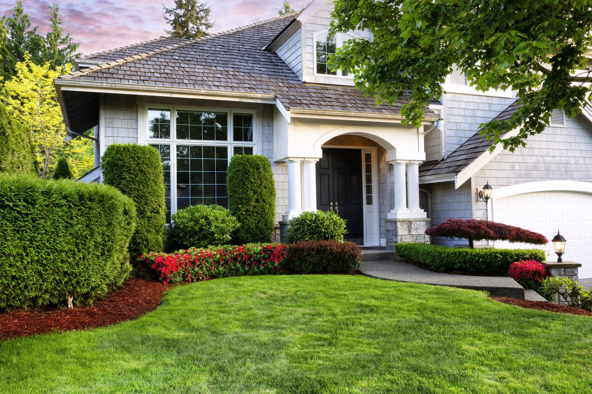 A two-story residential house with a well-manicured garden, green lawn, and a clear sky visible at dusk. The exterior features a white garage door and windows.