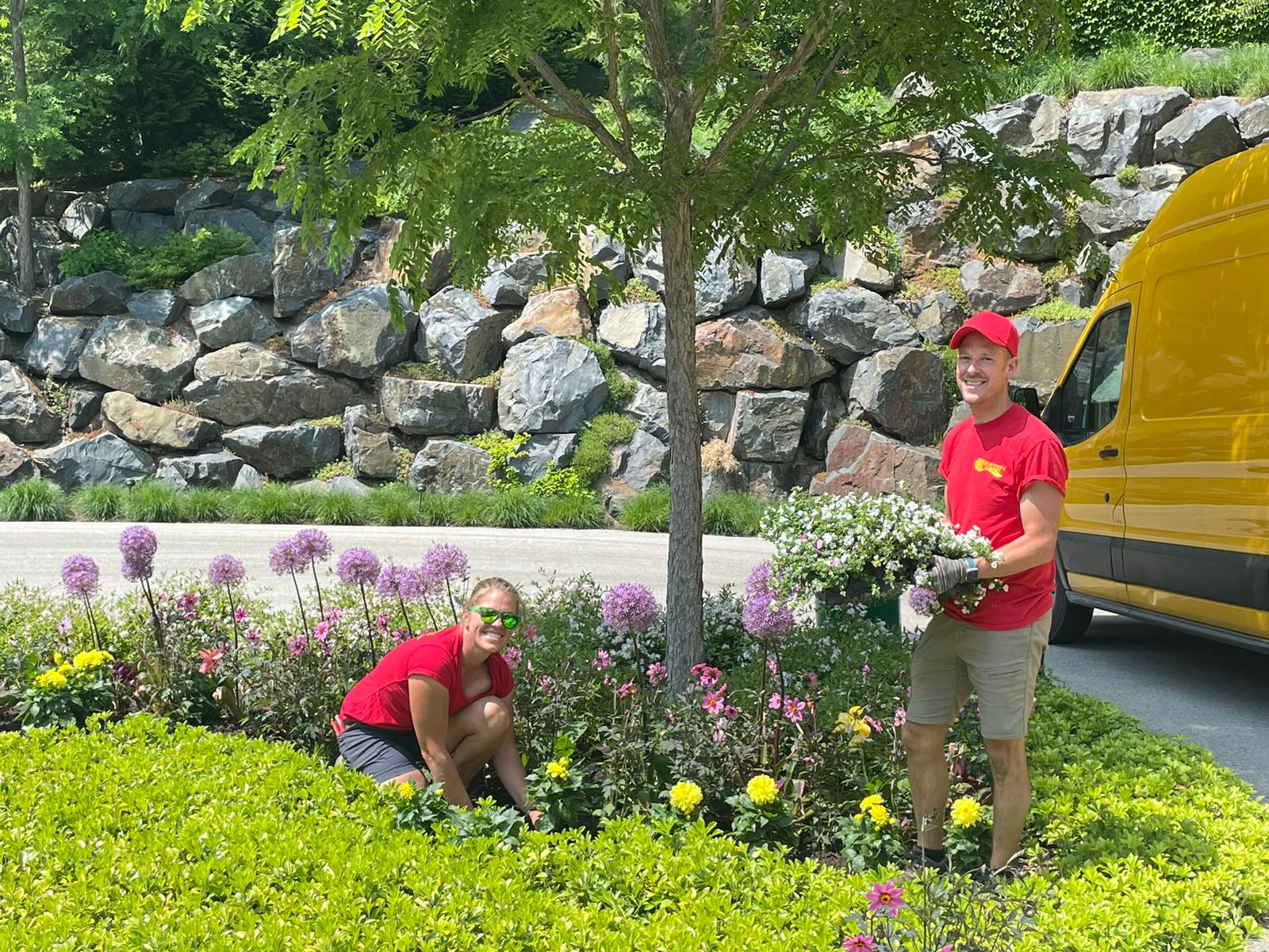 Two persons in red shirts and caps are gardening near a stone wall, with a yellow van parked nearby, surrounded by greenery and blooming flowers.