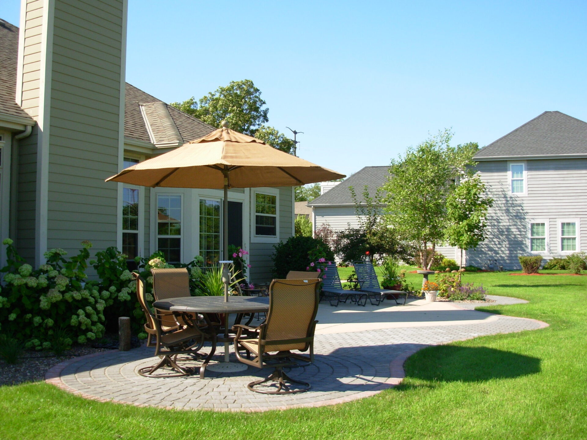 A sunny backyard with a patio dining set under an umbrella, surrounded by manicured grass, landscaped plants, and suburban homes.