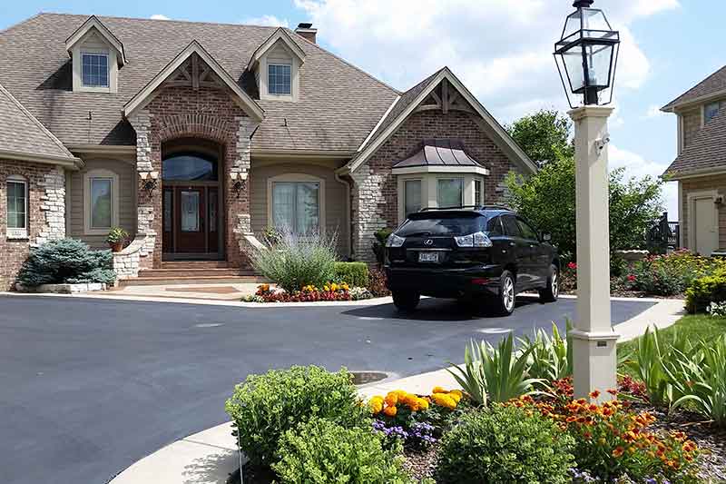 An upscale suburban home with a stone facade, landscaped garden, and a black SUV parked in the driveway under a clear sky. A street lamp stands in the foreground.