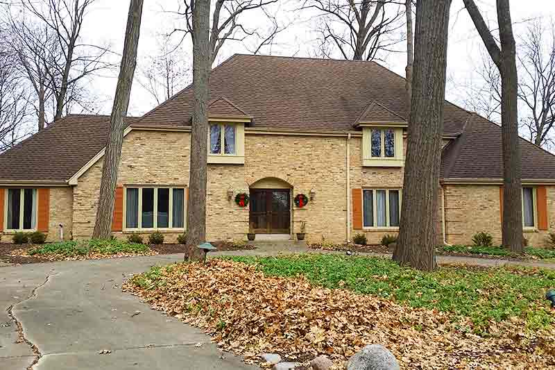 A two-story brick house with a brown roof, surrounded by trees and a lawn covered with fallen leaves. The front door has a holiday wreath.