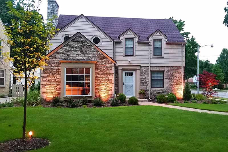 A two-story house with a well-manicured lawn at dusk. Warm lights illuminate the stone façade, and a young tree is lit by a ground light.