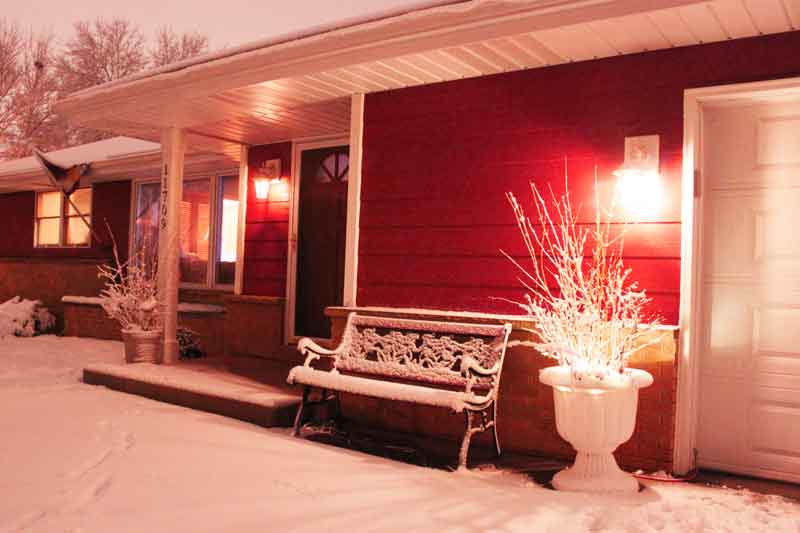 This image shows a cozy house exterior at twilight with red walls, snow-covered bench, porch, and plants after a winter snowfall. Warm lights glow warmly from windows.