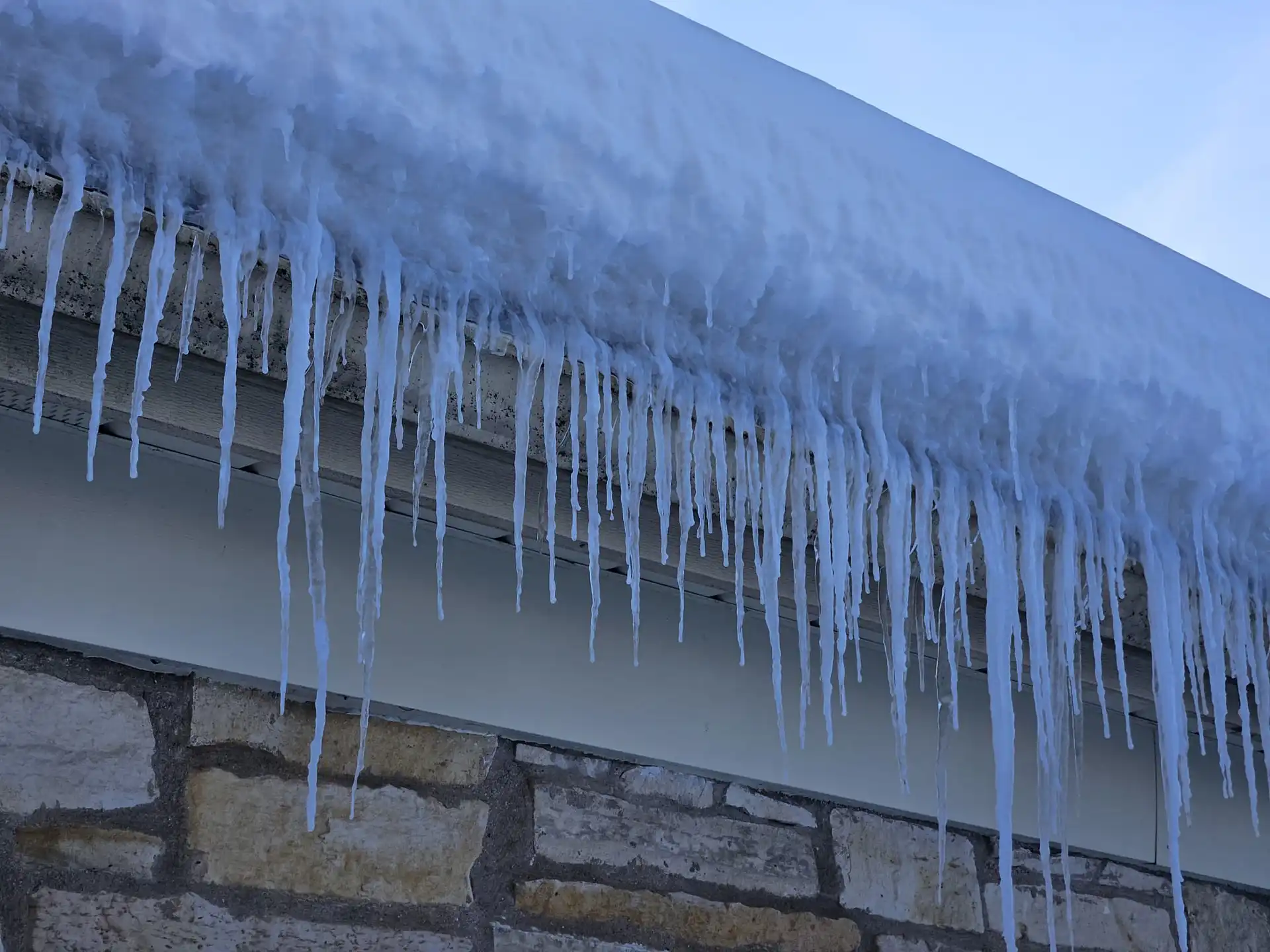A roof covered in snow with numerous long icicles hanging from the eaves above a stone wall, indicating cold winter conditions.