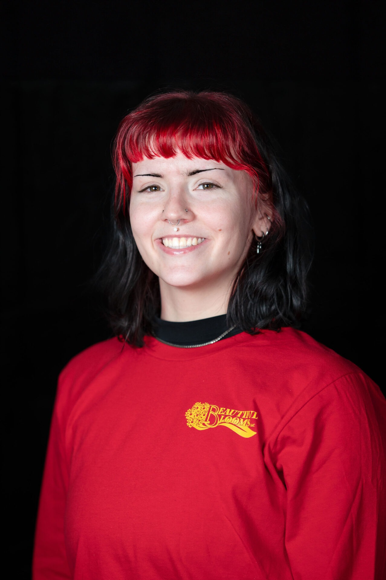 A person with bright red hair and a nose piercing is smiling, wearing a red shirt with "Beautiful Blooms" printed on it, against a black background.