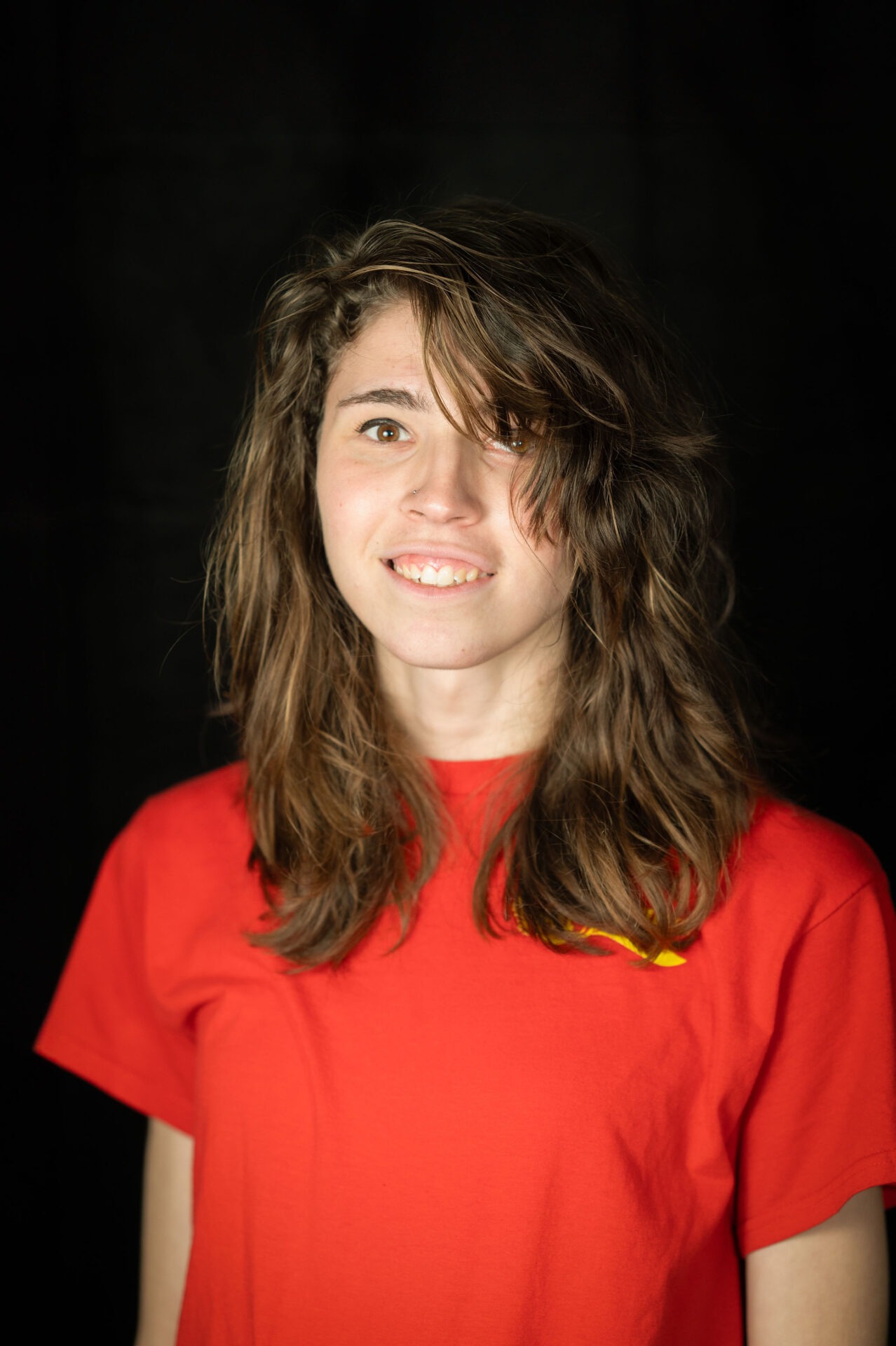 The image shows a smiling person with medium-length wavy hair, wearing a red shirt, in front of a dark background with soft lighting.