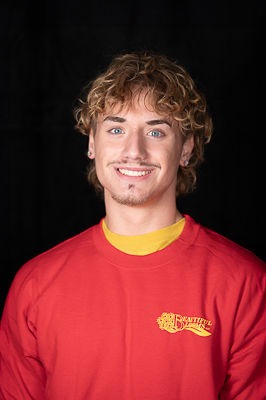 A person with curly hair is smiling against a black background, wearing a red shirt with a small logo and a yellow collar showing through.
