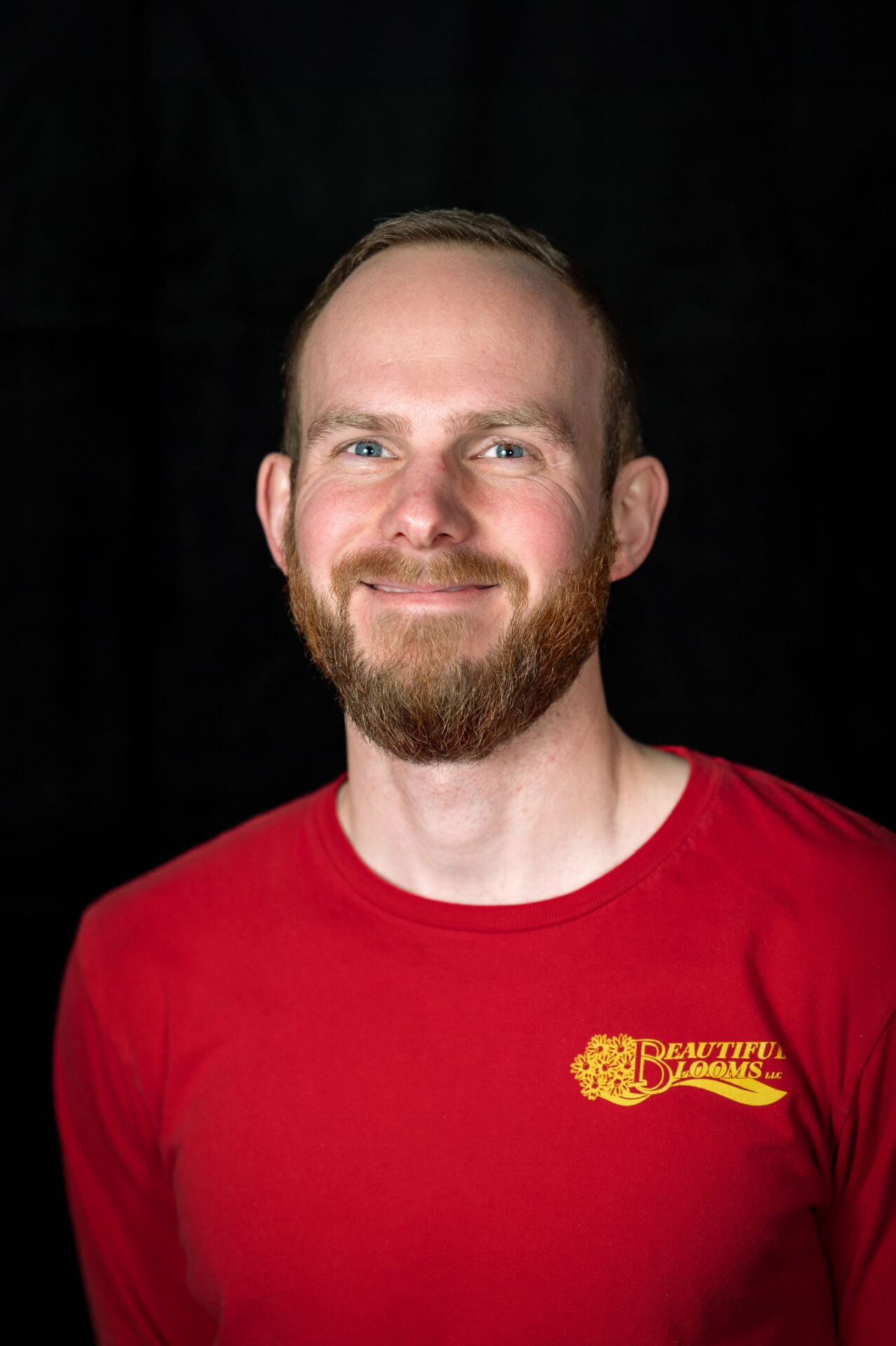 A person with a beard smiles warmly, wearing a red t-shirt with yellow text, set against a dark background in a portrait-style photograph.