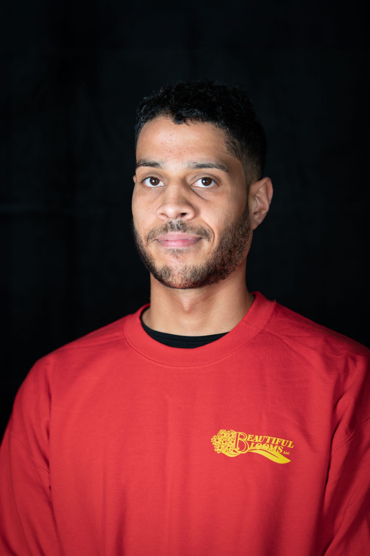 A person with short hair stands against a black backdrop, wearing a red t-shirt with "Beautiful Blooms" printed on it, looking directly at the camera.
