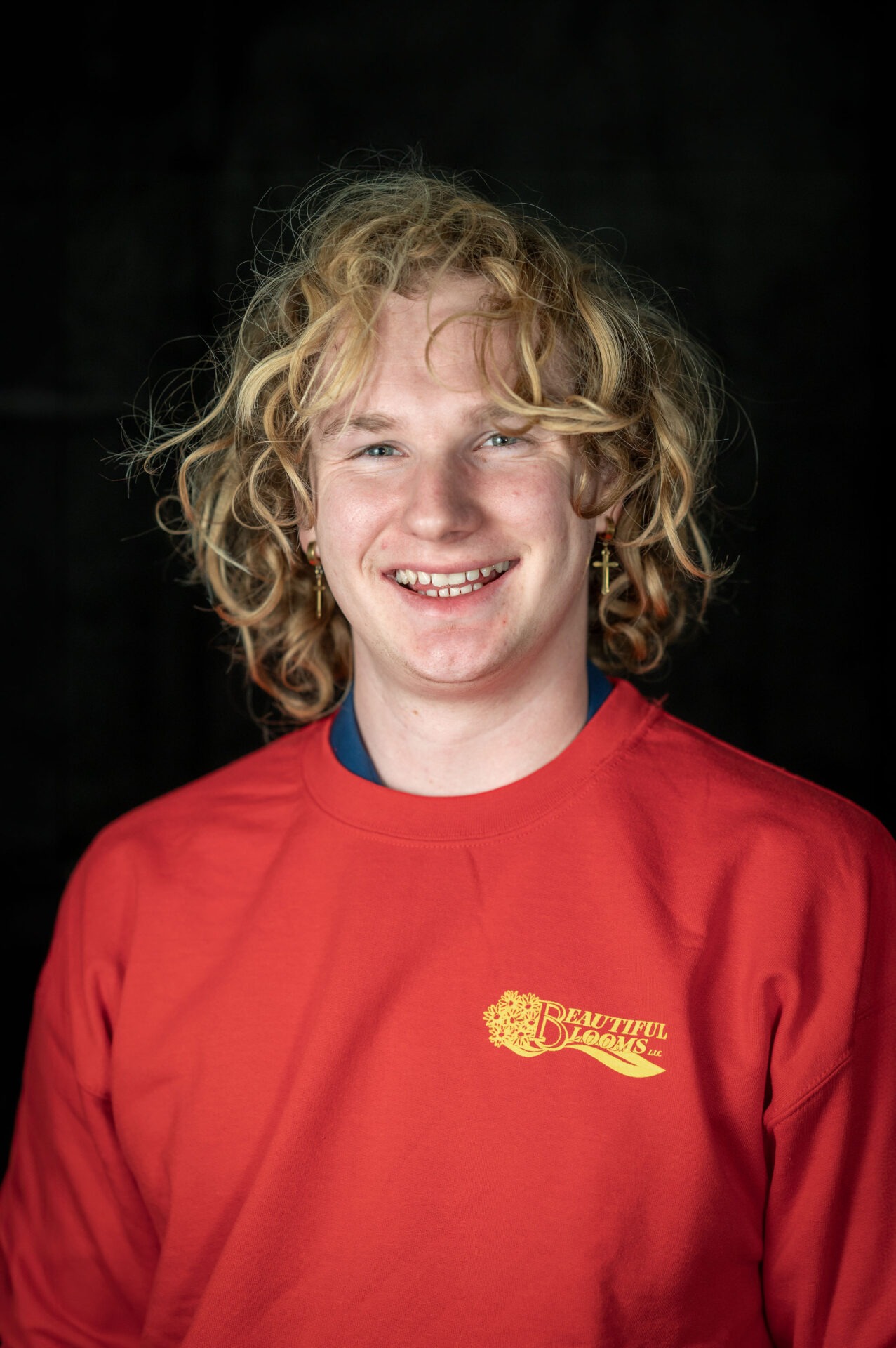 A smiling person with curly blonde hair, wearing a red long-sleeved shirt with a logo, against a dark background. The person exudes a joyful demeanor.