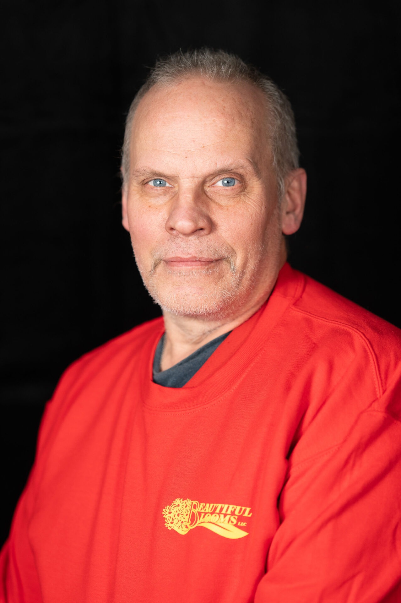 A person with light complexion and short hair, wearing a red shirt with a logo, stands before a black background, offering a faint smile.