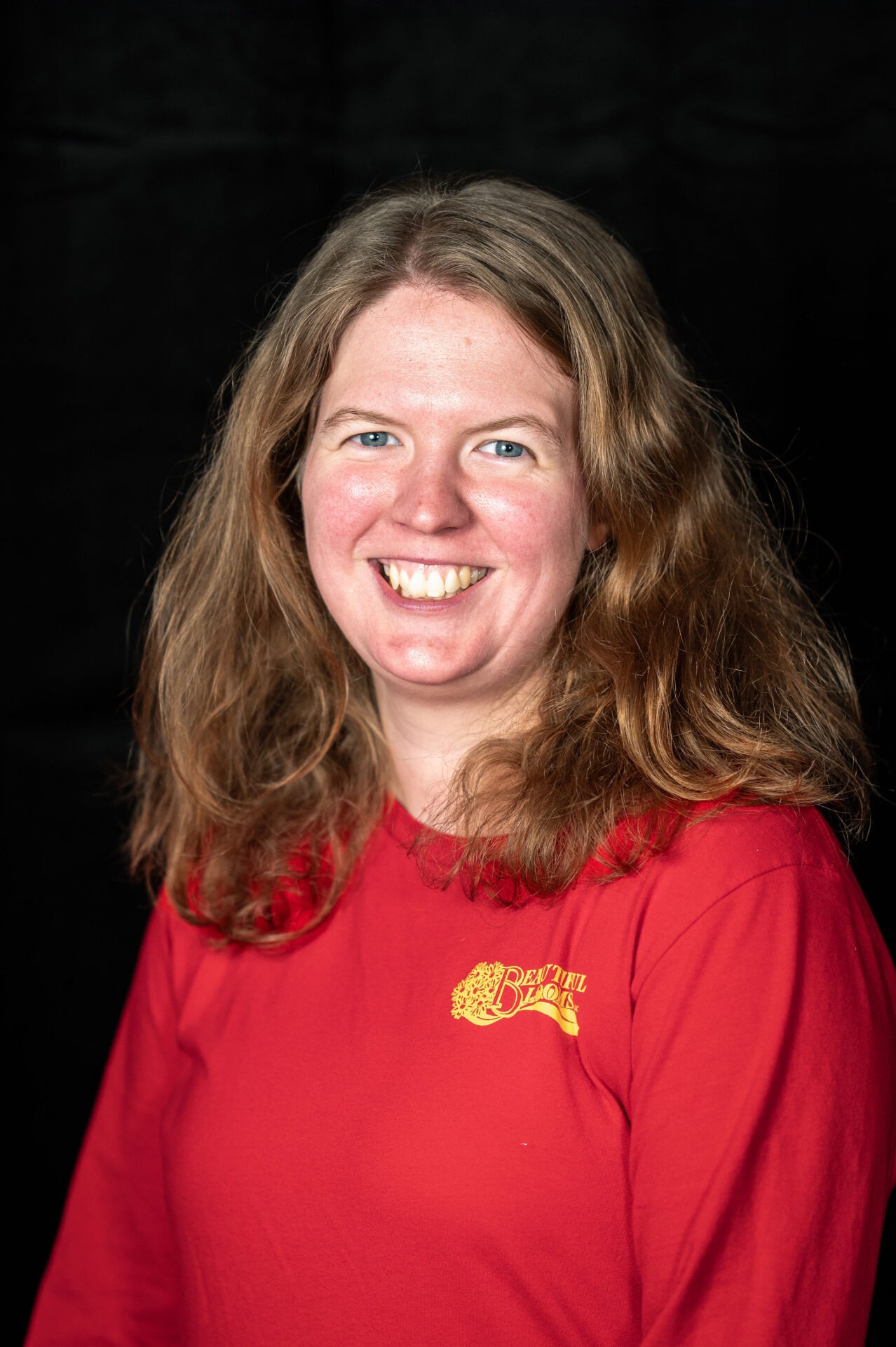 A smiling person with long blond hair wearing a red shirt with a "Beautiful Blonde" logo in front of a black background.