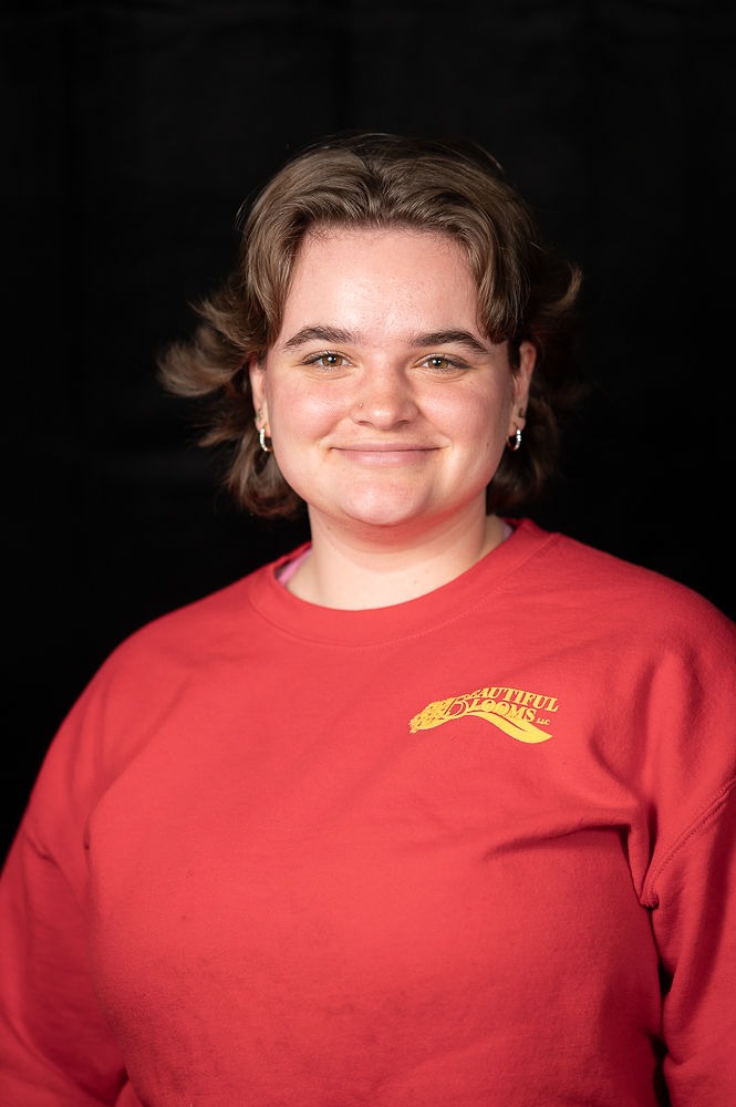 A person is smiling at the camera wearing a red shirt with yellow text. They have short hair, stud earrings, and a black background is behind them.