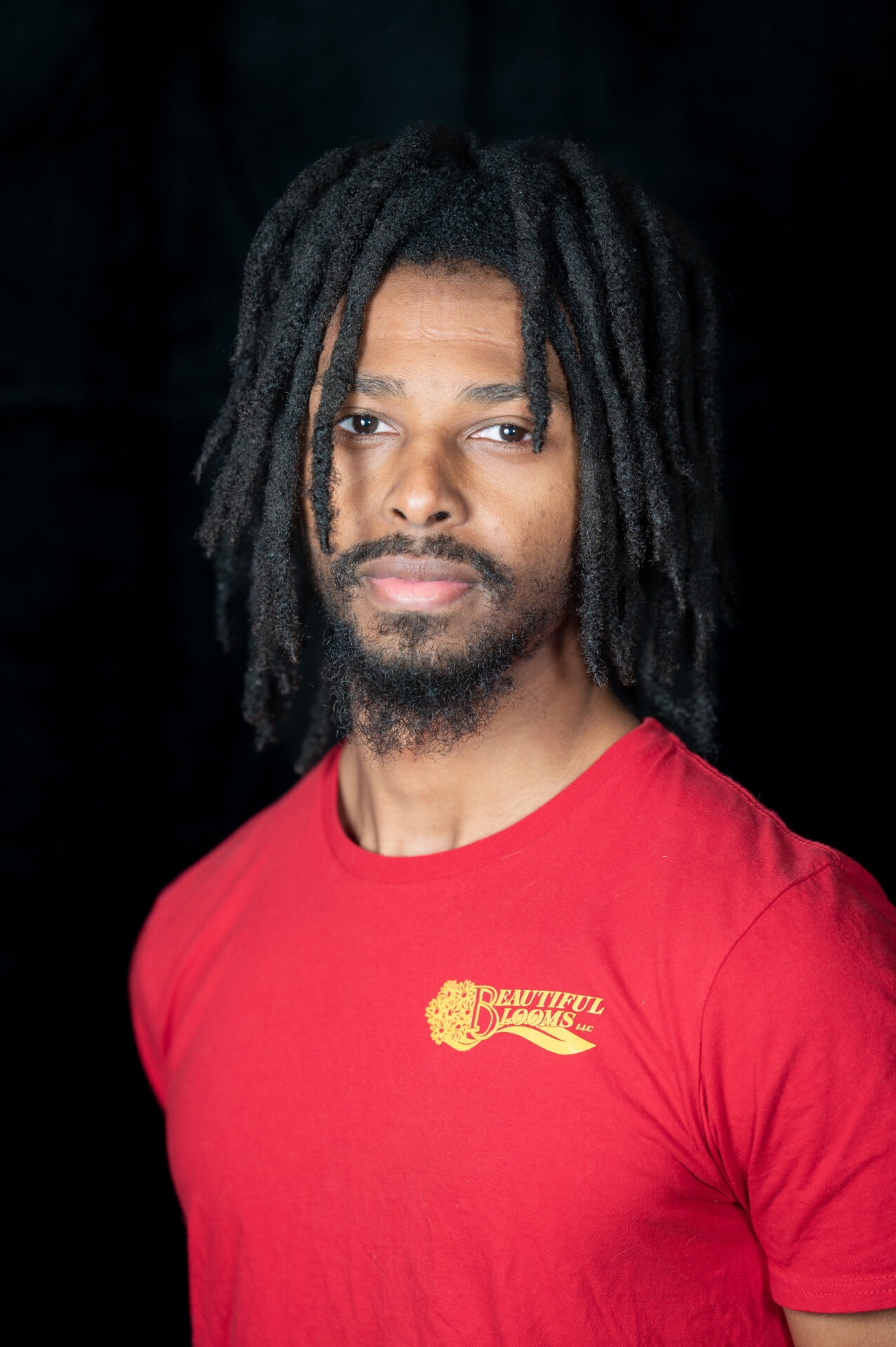 A person with shoulder-length dreadlocks is wearing a red t-shirt and looking calmly at the camera against a dark background.