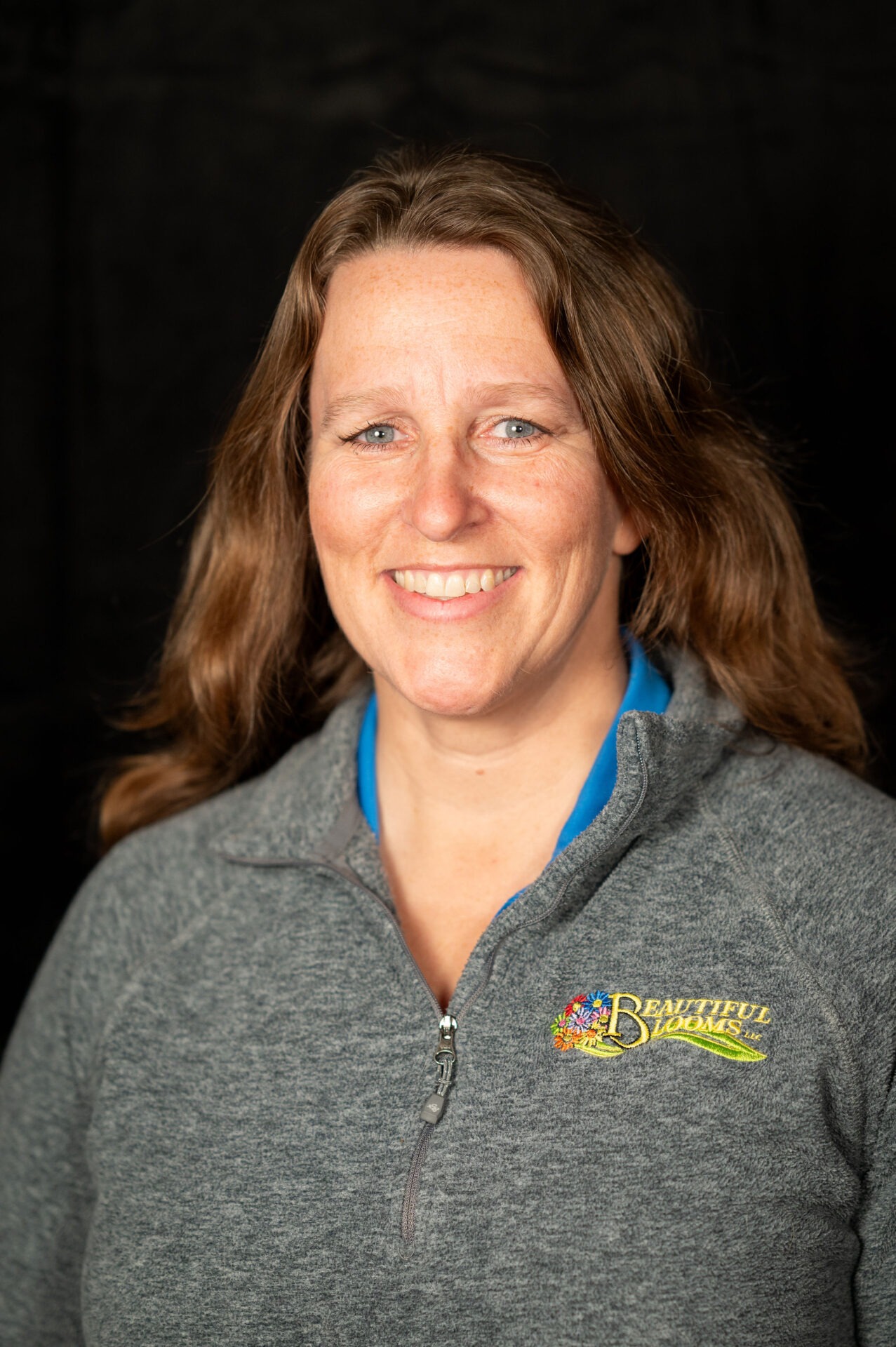 A smiling person is photographed against a black background. They are wearing a zipped grey fleece with an embroidered logo "Beautiful Blooms."