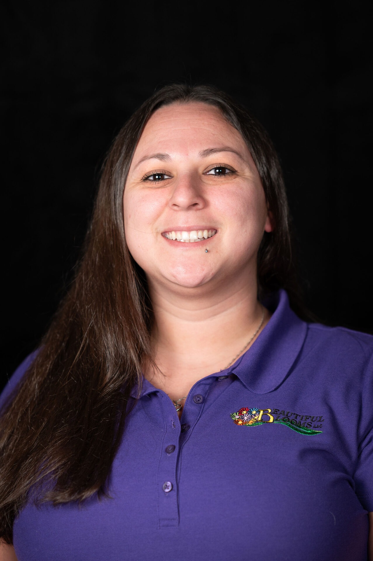 A smiling person with long brown hair is wearing a purple polo shirt with an embroidered logo on a black background. They exude a friendly demeanor.