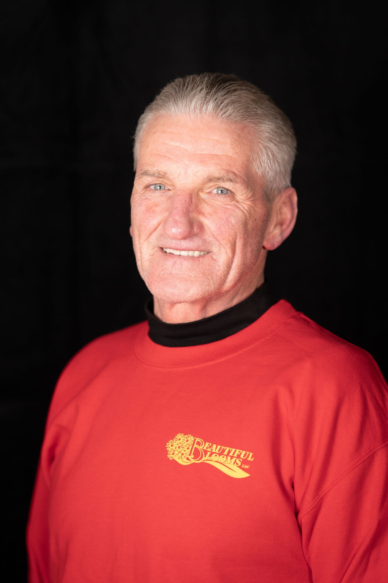 A smiling person with silver hair wearing a red turtleneck with a "Beautiful Blooms LLC" logo. The background is dark and unadorned.