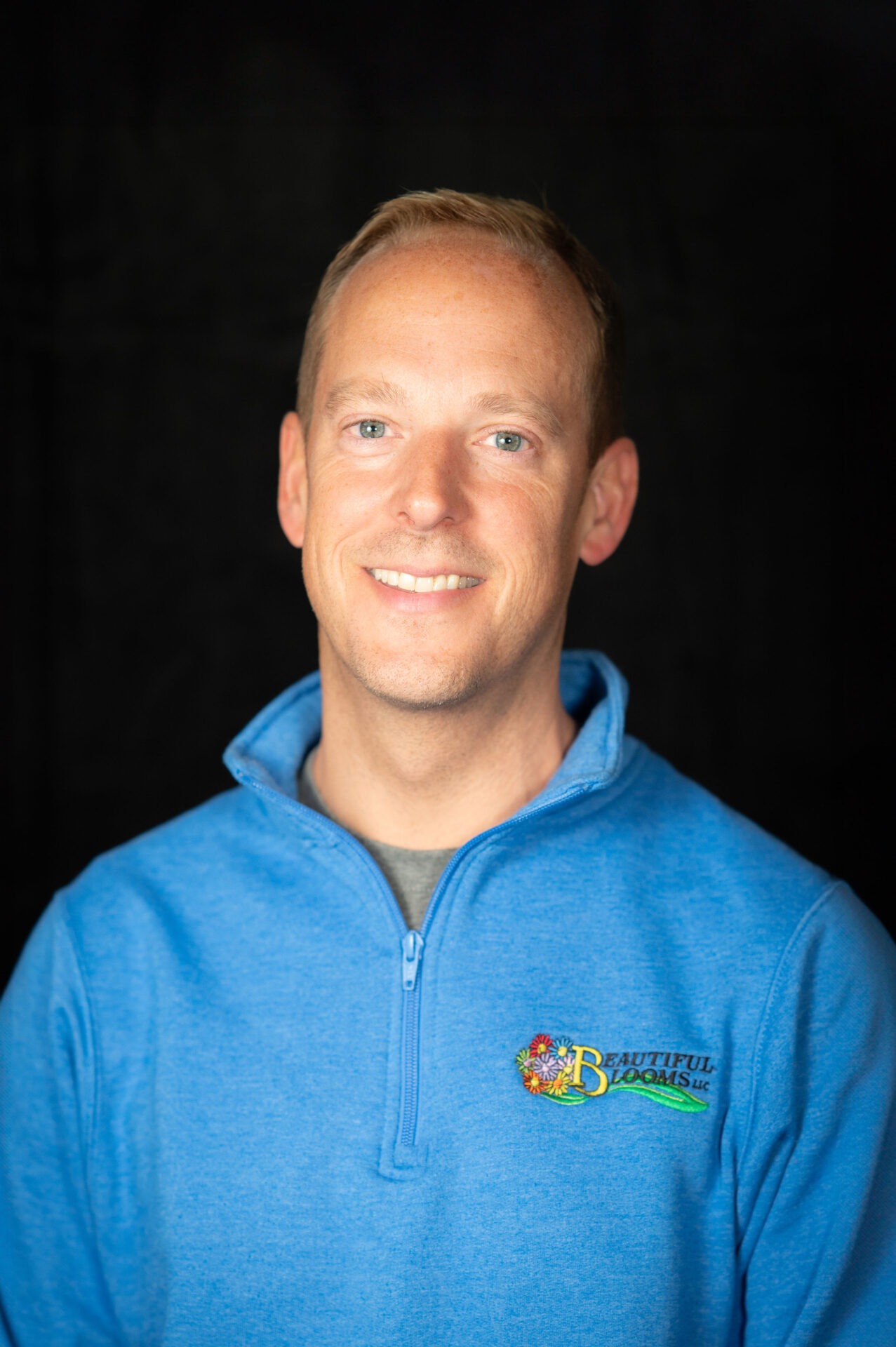 A person with short blonde hair and a light smile, wearing a blue pullover with a logo, poses against a black background.