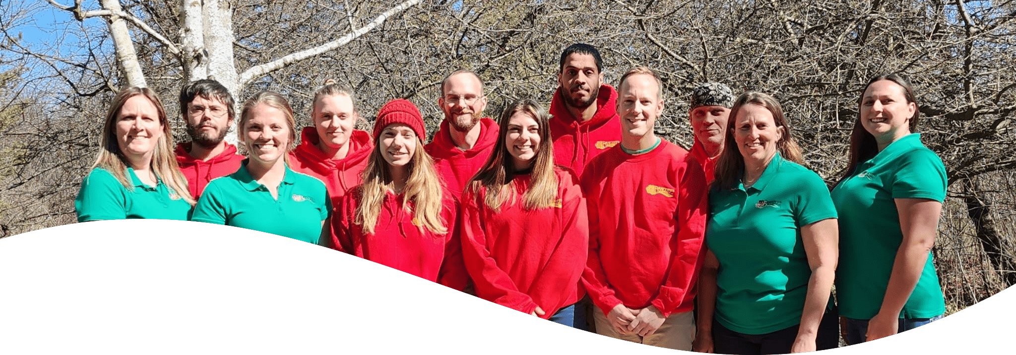 A group of eleven people standing outdoors, wearing green and red uniforms, smiling for a photo with a leafless tree background.