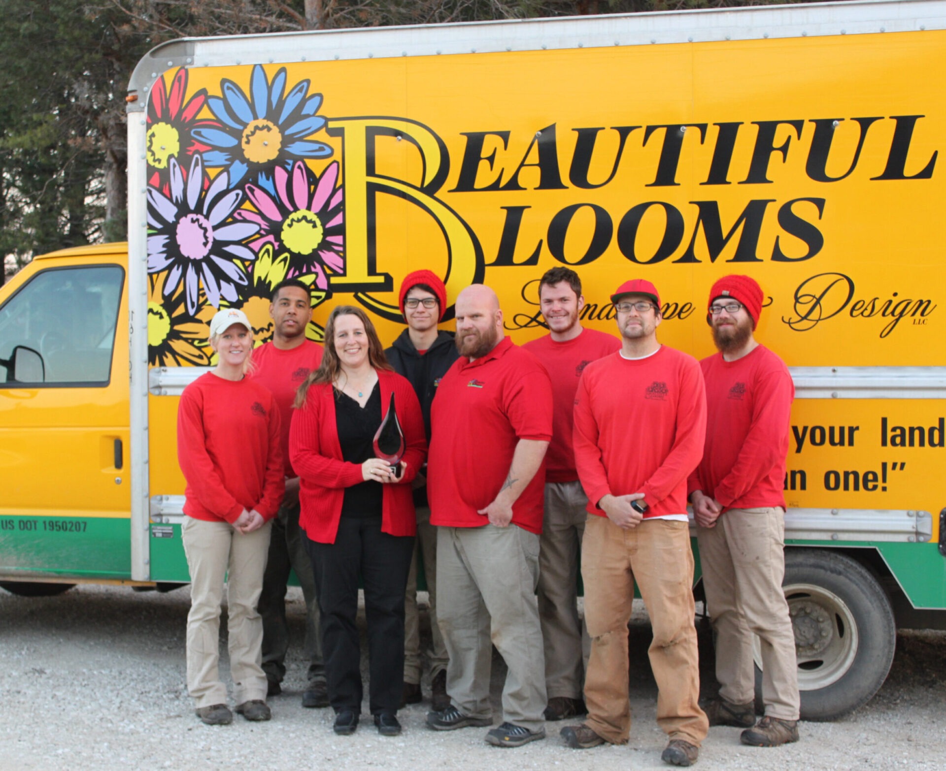 A group of eight people wearing red shirts stands in front of a yellow truck decorated with flowers and the words "Beautiful Blooms Landscape & Design."