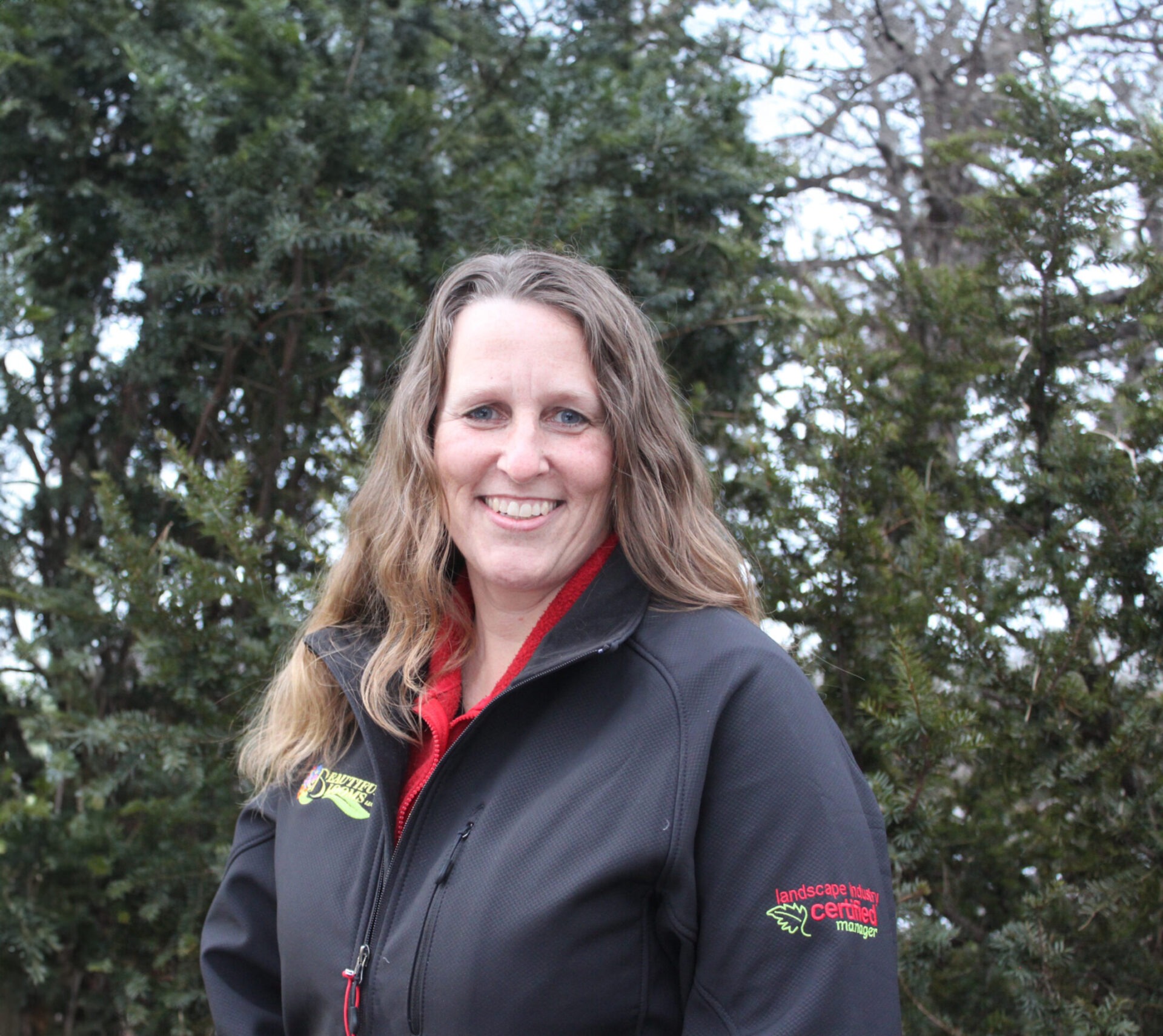 A smiling person with long hair stands in front of evergreen trees, wearing a black jacket with red detailing and an embroidered logo.