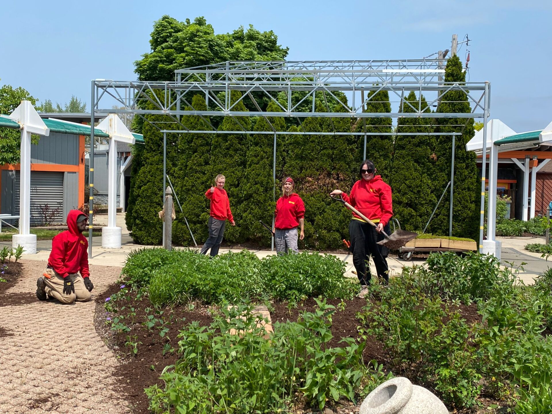 Five people in red shirts are engaged in gardening tasks, planting and maintaining a landscaped area with a metal structure in the background.