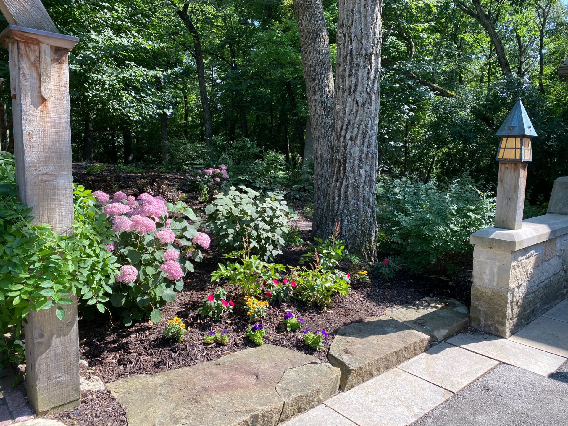 A peaceful garden with pink hydrangeas, assorted flowers, trees, a stepping stone path, and wooden and stone pillars with a lantern.